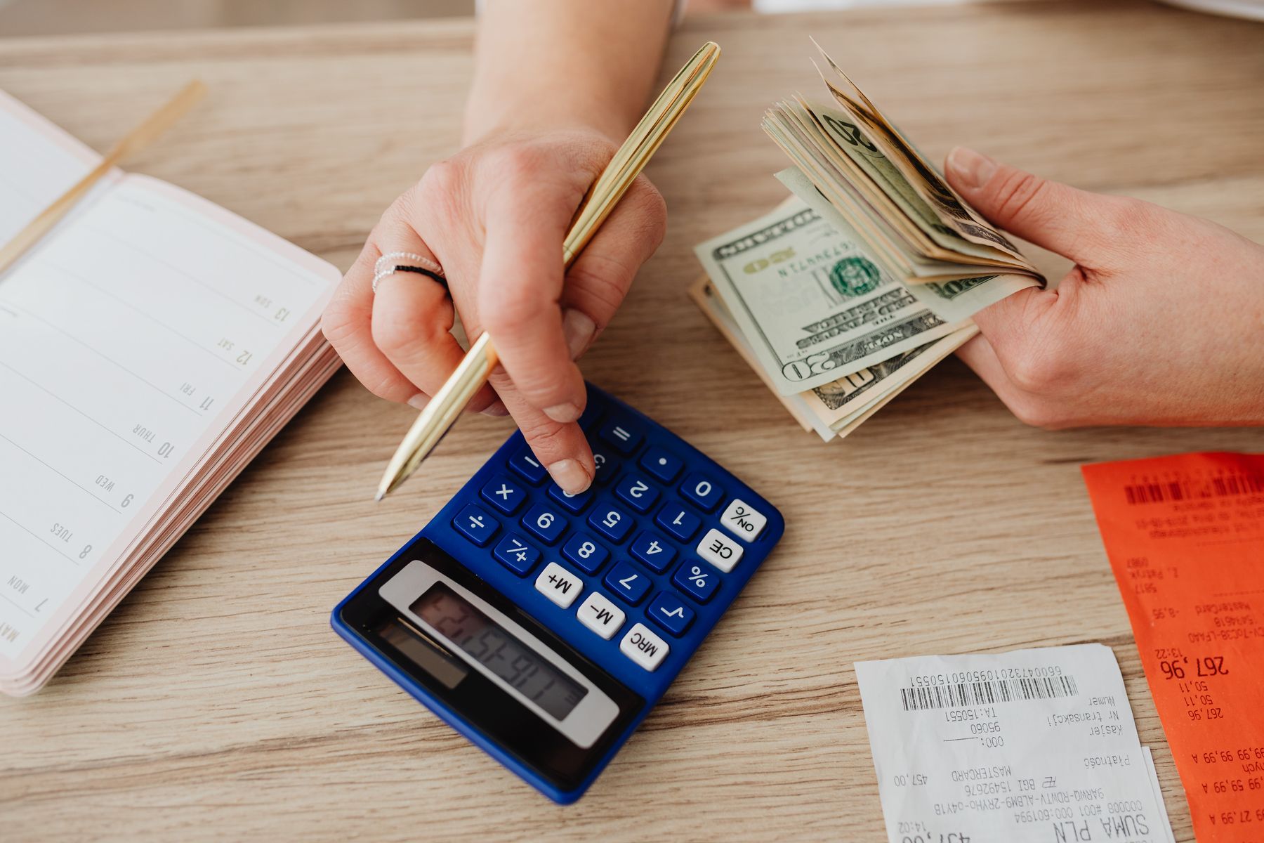Hands using a calculator beside a planner, receipts and cash while working through a household budget